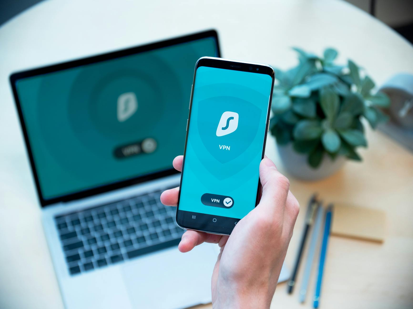 Smartfon z aplikacją VPN w dłoni na tle laptopa, symbol szyfrowania danych